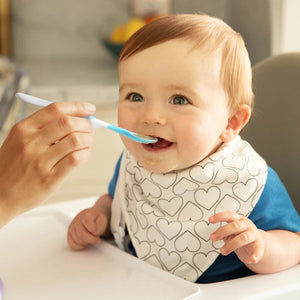 Baby being fed with a spoon by an adult, wearing a bib with heart patterns.