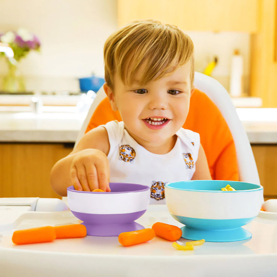 Child sitting in a high chair with two colorful bowls and carrots on a kitchen counter.